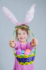 Little girl wearing pink shirt and easter bunny ears. She holds holds a basket of easter eggs in front of camera.  