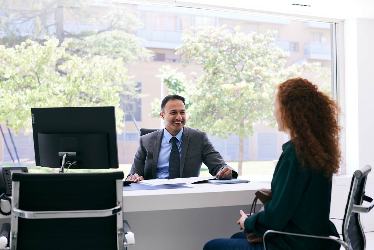 A man and a woman sit at a desk across from each other talking.